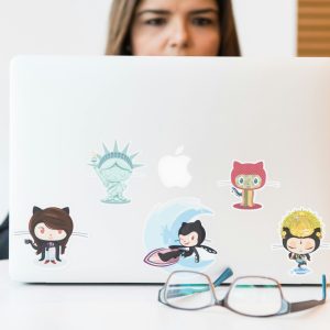 Woman focused on laptop with colorful GitHub-themed stickers, eyeglasses in foreground.