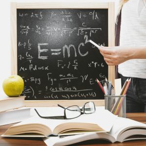 Woman writing physics equations on a blackboard with books and an apple on the desk.